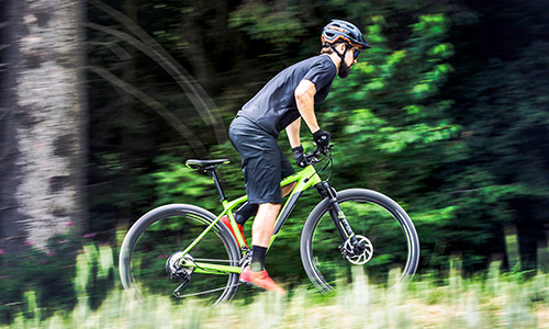 Pyöräilijä ajaa vihreällä maastopyörän hardtail-pyörällä seisoma-asennossa vihreässä luonnonympäristössä.