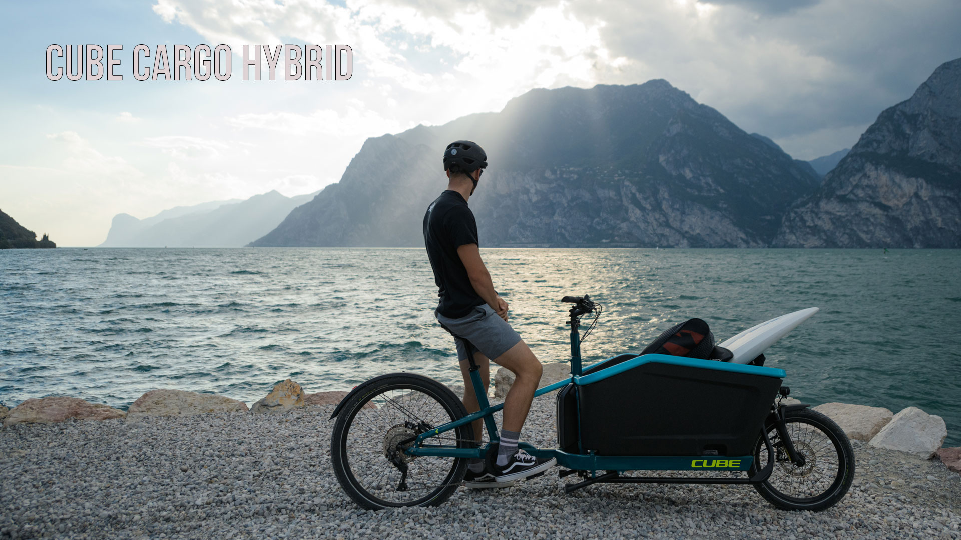 De foto toont een man op een blauwe bakfiets die aan de oever van een meer staat en naar de bergen op de achtergrond kijkt. De foto is gelabeld met de titel Cube Cargo Hybrid.